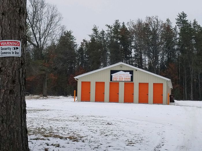 Warning sign on a pine tree reading security cameras in use, with a storage building behind it
