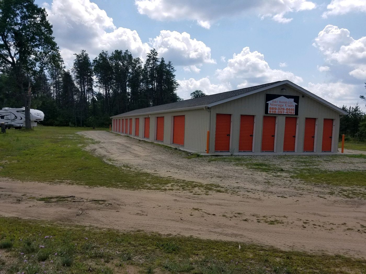 Long tan storage building with a row of orange roll-up doors, a gravel drive and pine trees behind it